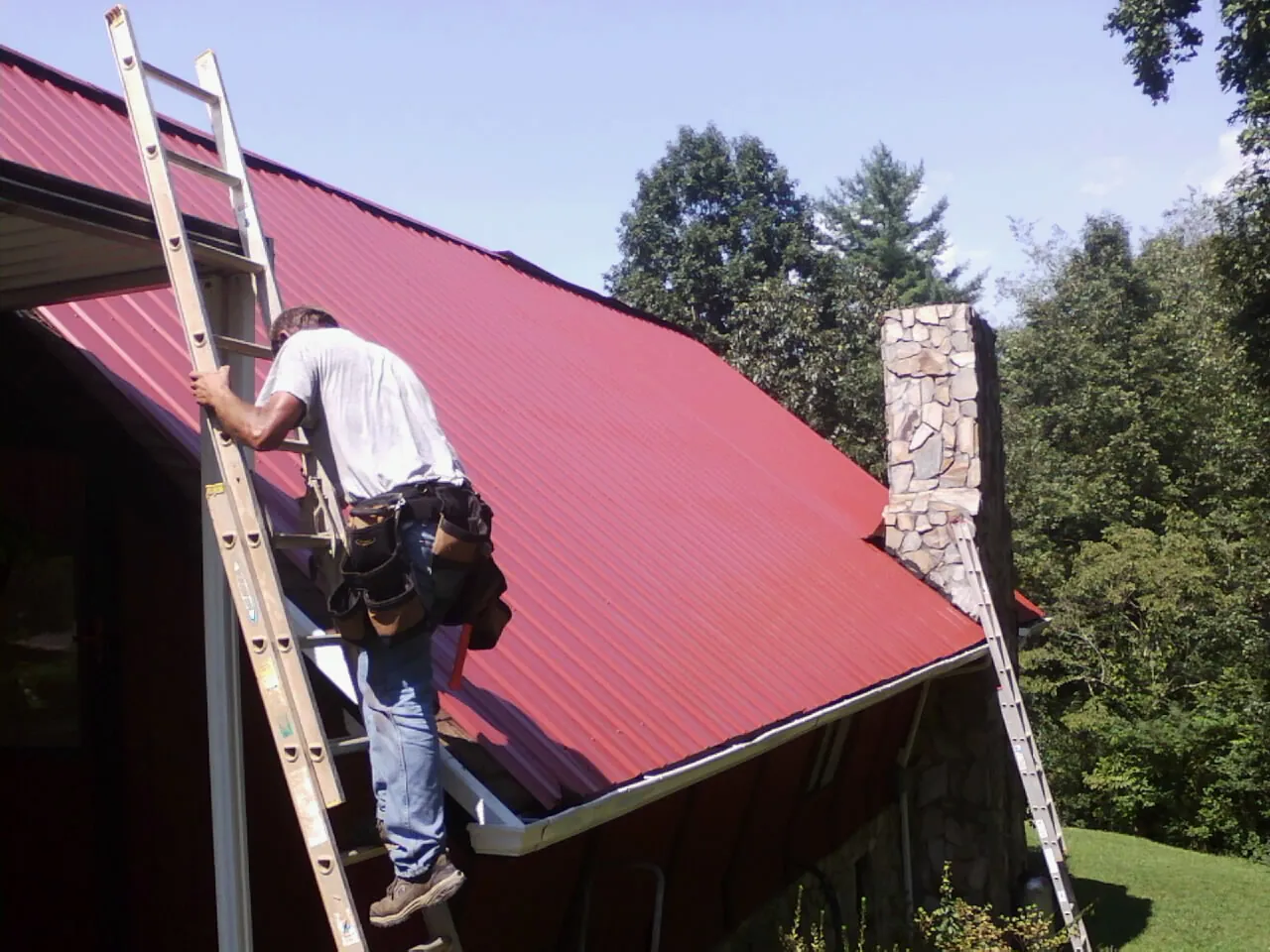 Roofer on a ladder beside a red metal roof and stone chimney — Soffit & Fascia Repair by Roofing Brothers in Lee Center